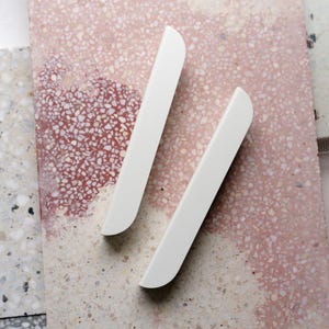 Peut inclure: Deux poignées de meubles blanches et incurvées sont positionnées en diagonale sur une surface rose et blanche mouchetée. Les poignées ont une finition lisse et mate et un design minimaliste. L'arrière-plan présente un motif de type terrazzo.