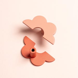 Puede incluir: Dos tiradores de armario con forma de nube en tonos coral y melocotón. Uno está completo, el otro desmontado, mostrando los componentes de fijación. Los tiradores metálicos tienen un acabado mate. El fondo es rosa suave.