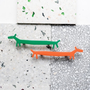 Könnte beinhalten: Zwei Dackel-förmige Schrankgriffe, einer grün und einer orange, werden auf einer gesprenkelten Oberfläche präsentiert. Die Griffe bestehen aus einem festen Material und haben ein minimalistisches Design. Der Hintergrund zeigt verschiedene Terrazzofliesen.