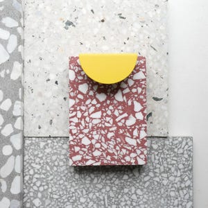 Peut inclure: Un objet rectangulaire, moucheté de rouge et de blanc, surmonté d'un demi-cercle jaune. L'objet est posé sur un fond de surfaces mouchetées grises et blanches, créant une esthétique moderne et minimaliste.