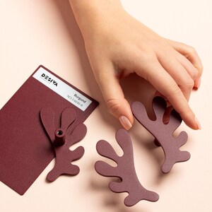 K&ouml;nnte beinhalten: Burgunderfarbene Schrankbeschl&auml;ge mit einem einzigartigen, organischen Design. Die Beschl&auml;ge haben die Form einer stilisierten Pflanze oder Koralle. Ein Farbmuster mit dem Text "DESIVA" und "Burgund" ist ebenfalls vorhanden.