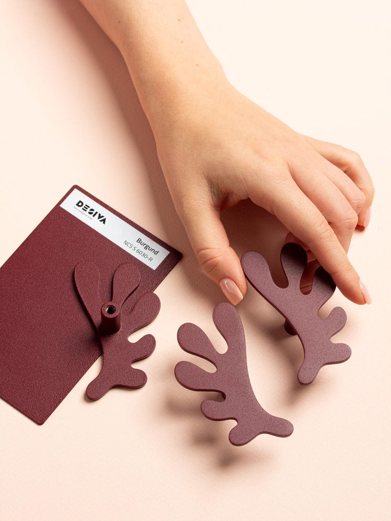 K&ouml;nnte beinhalten: Burgunderfarbene Schrankbeschl&auml;ge mit einem einzigartigen, organischen Design. Die Beschl&auml;ge haben die Form einer stilisierten Pflanze oder Koralle. Ein Farbmuster mit dem Text "DESIVA" und "Burgund" ist ebenfalls vorhanden.