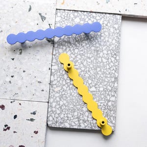 Könnte beinhalten: Zwei wellenförmige Schrankgriffe, einer in Periwinkleblau und einer in Gelb, werden auf einer gesprenkelten grauen Oberfläche gezeigt. Die Griffe haben ein gewelltes Kantendesign und sind aus Metall gefertigt. Die Schrankbeschläge sind ein modernes Design.