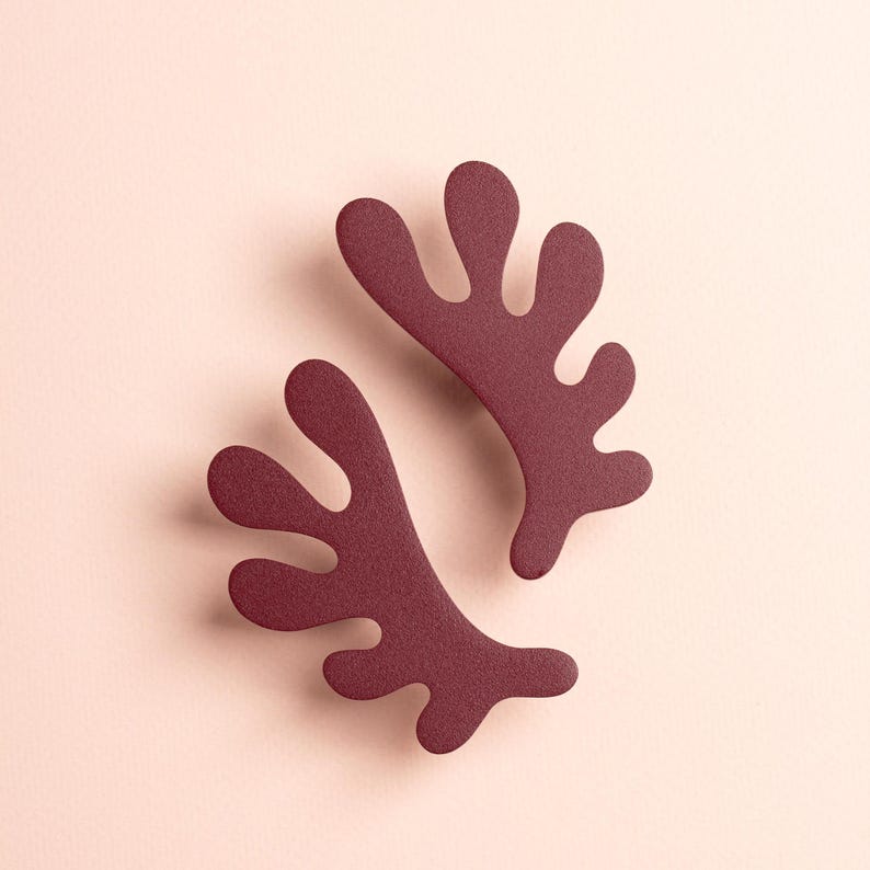 K&ouml;nnte beinhalten: Zwei weinrote, abstrakte, blattf&ouml;rmige Dekorationsgegenst&auml;nde. Die Objekte haben eine strukturierte Oberfl&auml;che und sind auf einem hellrosa Hintergrund angeordnet. Das Design ist modern und minimalistisch, geeignet f&uuml;r Heimdekoration oder Kunstprojekte.