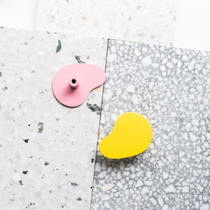Könnte beinhalten: Nahaufnahme von zwei abstrakt geformten Objekten. Eines ist hellrosa mit einem kleinen zylindrischen Vorsprung, das andere leuchtend gelb. Sie liegen auf einer Oberfläche mit einem gesprenkelten Terrazzo-Muster.