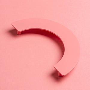 Puede incluir: Un tirador de armario semicircular de color rosa suave. El tirador presenta una forma curvada con dos pequeños postes cilíndricos en cada extremo para el montaje. El fondo es rosa a juego, creando una estética monocromática.
