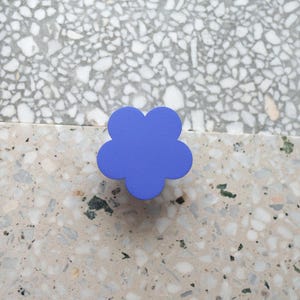 Può includere: Un pomello per armadio a forma di fiore blu. Il pomello è di un colore blu solido e vibrante ed è posto su una superficie maculata di colore chiaro. Il design è semplice e moderno, adatto a vari stili di mobili.