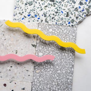 Może przedstawiać: Abstrakcyjna kompozycja z płytkami lastryko w odcieniach szarości, bieli i błękitu. Żółta i różowa falista linia umieszczone są na płytkach, tworząc nowoczesny i kolorowy design. Obraz ma czystą, minimalistyczną estetykę.