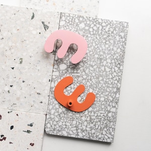 Peut inclure: Deux boutons de meuble abstraits. L'un est rose clair et l'autre orange. Les deux boutons ont un design à trois lobes. Les boutons sont posés sur un fond de carreaux mouchetés gris et blancs.