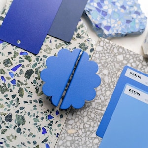 May include: A pair of blue, flower-shaped bookends. The bookends are on a surface with various samples of blue and grey materials, including paint swatches and terrazzo tiles. The word "DESIYA" is visible on the paint swatches.