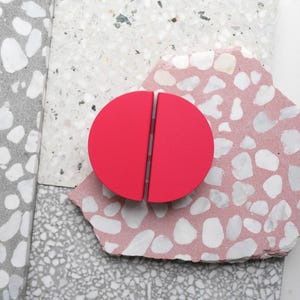 May include: A vibrant red, semi-circular cabinet pull, split in two, sits on a pink and white speckled stone. The pull contrasts with the neutral background of stone samples, showcasing a modern design. The cabinet pull is approximately 10 cm in diameter.