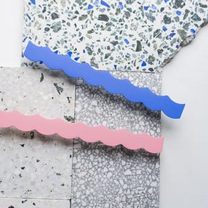 Könnte beinhalten: Eine Studioaufnahme zeigt zwei wellenförmige, dekorative Elemente in Blau und Rosa, die auf einer strukturierten, gesprenkelten Oberfläche platziert sind. Das blaue Element befindet sich über dem rosa Element. Der Hintergrund besteht aus verschiedenen Fliesen im Terrazzo-Stil.