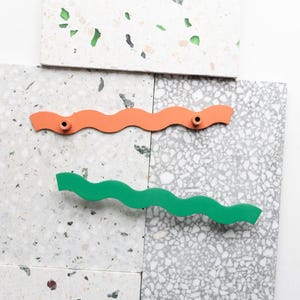 Könnte beinhalten: Zwei wellenförmige Schrankgriffe, einer orange und einer grün, werden vor einem Hintergrund aus gesprenkelten grau-weißen Fliesen gezeigt. Jeder Griff hat zwei Montagelöcher. Die Griffe sind etwa 25 cm lang.