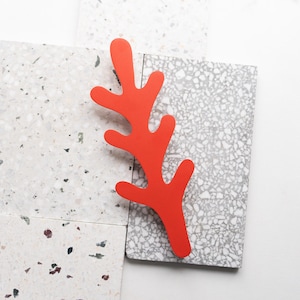 Op de afbeelding: Een feloranje, koraalvormig decoratief object ligt op een verzameling grijze en witte terrazzotegels. Het koraal heeft een levendige, effen kleur en de tegels hebben een gespikkeld patroon. De compositie is eenvoudig en modern.