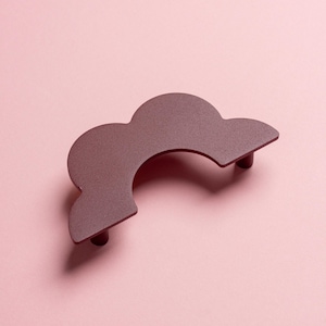 May include: A burgundy-colored, cloud-shaped cabinet pull with two small cylindrical posts. The hardware is made of metal and has a matte finish. The pull is set against a light pink background.