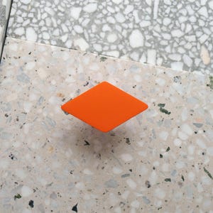 Könnte beinhalten: Ein orangefarbener, rautenförmiger Schrankgriff. Die Hardware befindet sich auf einer gesprenkelten, hellfarbigen Oberfläche. Der Griff ist ein einfaches, geometrisches Design, geeignet für Schubladen oder Schränke.