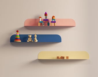 Könnte beinhalten: Drei wandmontierte schwebende Regale in Pink, Blau und Gelb mit abgerundeten Enden. Das pinke Regal trägt bunte Holzbausteine. Das blaue Regal zeigt ein Stapelring-Spielzeug, eine Bärenfigur und einen Hasen. Das gelbe Regal präsentiert Holzklötze, die das Wort TIDYS bilden.