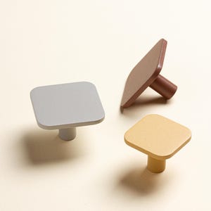 Op de afbeelding: Drie vierkante kastknoppen in verschillende kleuren. Eén is lichtgrijs, één is bruin en de andere is geel. Elke knop heeft een vierkant vlak en een cilindrische steel. De knoppen zijn gemaakt van hout of een vergelijkbaar materiaal.
