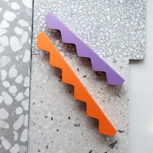 Può includere: Due maniglie per mobili a zig-zag, una arancione e una lilla, su uno sfondo di pietra screziata grigia e bianca. Le maniglie hanno un design moderno e minimalista. Le maniglie sono lunghe circa 20 cm.