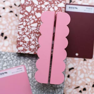 Peut inclure: Deux serre-livres roses à bords festonnés. Ils sont placés verticalement, avec un espace entre eux. En arrière-plan, des carreaux texturés roses, bruns et gris, ainsi que des nuanciers de couleurs.