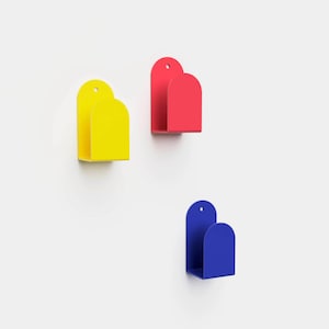 Peut inclure: Trois poches de rangement murales aux couleurs primaires vives. Une poche jaune, une rouge et une bleue, chacune présentant un design en forme d'arche arrondie avec un petit trou circulaire en haut pour la fixation. Les poches sont ouvertes.