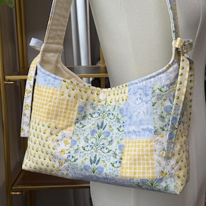 May include: A quilted shoulder bag with a patchwork design. The bag features a combination of floral, gingham, and patterned fabrics in shades of yellow, blue, and white. The strap is a solid beige color, and the bag is fastened with a white button.