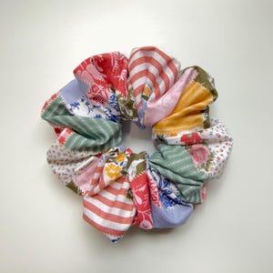 May include: A colorful scrunchie made from patchwork fabric. The scrunchie features various patterns, including floral, striped, and solid designs in red, pink, blue, green, and yellow. This hair accessory is a stylish addition to any hairstyle.