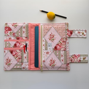 May include: Open quilted journal cover with a floral patchwork design in pink, red, and green. Features a gold zipper, interior pockets, and a strap with snap closures. Includes a pencil and a lemon.