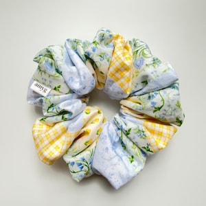 May include: A floral scrunchie with a mix of patterns, including blue floral, yellow gingham, and cream-colored fabric. The scrunchie has a tag that reads "amyg".
