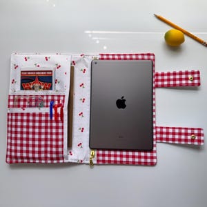 May include: A red and white gingham iPad case with a cherry print interior. The case has pockets for pens, a notepad, and a small card. A pencil and a yellow fruit are on the white surface.