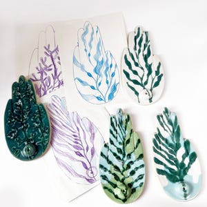 Könnte beinhalten: Handförmige Keramik-Räucherstäbchenhalter in Grün- und Blautönen mit botanischem Design. Die Halter werden mit Skizzen des Designs präsentiert. Die Räucherstäbchenhalter sind ein einzigartiger Einrichtungsgegenstand.