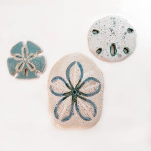Pode incluir: Três decorações de cerâmica em forma de dólar de areia em tamanhos variados. Os dólares de areia são brancos com detalhes em azul e verde-azulado. O maior tem um desenho floral no centro.