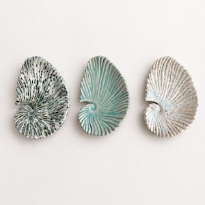 Könnte beinhalten: Drei keramische Muschelschalen in einer Reihe. Die Schalen sind in Blau-, Grau- und Weißtönen gehalten. Jede Schale hat ein Spiralmuster und eine strukturierte Oberfläche. Die Schalen werden vor einem weißen Hintergrund präsentiert.