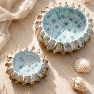 Pode incluir: Duas tigelas de cerâmica com um design inspirado no mar. As tigelas têm um interior azul claro com um padrão salpicado e uma borda texturizada em forma de sol com detalhes em castanho. Conchas marinhas espalhadas ao redor das tigelas em uma superfície de madeira clara.