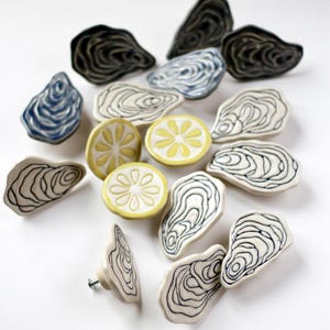 Könnte beinhalten: Eine Sammlung von Keramik-Schrankknöpfen in verschiedenen Formen, darunter Austernschalen und Zitronenscheiben. Die Knöpfe sind hauptsächlich weiß mit schwarzen, blauen und gelben Akzenten. Einige haben eine Schraube zur Befestigung.