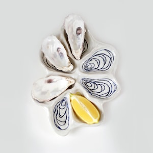 Könnte beinhalten: Eine weiße Austernservierplatte aus Keramik mit sechs Fächern, die jeweils mit einem marineblauen Design umrandet sind. Die Platte enthält vier geöffnete Austern und eine Zitronenspalte. Die Austern haben ein perlmuttartiges Inneres.