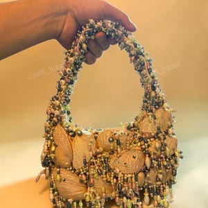May include: A handbag constructed from numerous tan seashells, featuring a handle and body densely embellished with a variety of multi-coloured beads in shades of green, pink, and metallic grey, with dangling beaded fringe accents throughout.