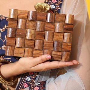 Op de afbeelding: Een handgemaakte houten clutch met een geometrisch patroon. De clutch is gemaakt van kleine, rechthoekige houten blokken gerangschikt in een raster. Het heeft een goudkleurige bloemen sluiting. De algehele kleur is een warm bruin.