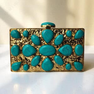 Könnte beinhalten: Eine rechteckige, goldfarbene Clutch mit einer stark strukturierten, zerknitterten Metalloberfläche. Die Vorderseite ist mit zahlreichen glatten, tropfenförmigen und ovalen türkisfarbenen Steinen mit dunkler Maserung verziert. Ein einzelner Türkis dient als Verschluss.