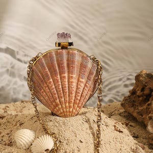 May include: A seashell-shaped clutch bag with a gold chain strap. The bag is made from a large, fan-shaped shell with a natural, textured surface. It is adorned with a gold-coloured metal frame and a purple crystal clasp. Two small white seashells are in the sand.