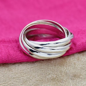 Op de afbeelding: Een zilverkleurige ring met vijf in elkaar grijpende banden. De ring ligt op een helder roze stof. De banden zijn glad en afgerond, waardoor het licht wordt gereflecteerd.