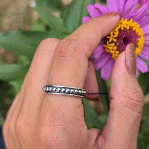 Peut inclure: Une bague en argent avec un motif de perles est présentée sur un doigt. La bague est mise au point, avec un arrière-plan flou d'une fleur violette et de feuilles vertes. La surface de la bague est réfléchissante, et les perles sont espacées uniformément.