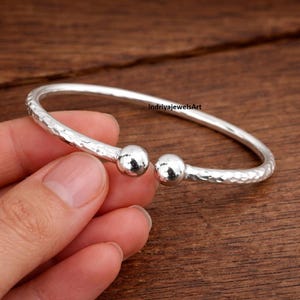 Op de afbeelding: Een zilveren armband met een gehamerde textuur en twee bolvormige eindkappen. De armband wordt in een hand gehouden en de achtergrond is een houten oppervlak. De tekst "IndriyajewelsArt" is zichtbaar.