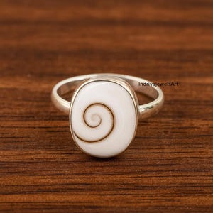 Puede incluir: Anillo de plata con una piedra ovalada blanca con un diseño de espiral marrón. El anillo tiene una banda de plata pulida y una superficie lisa. El anillo está sobre un fondo de madera.