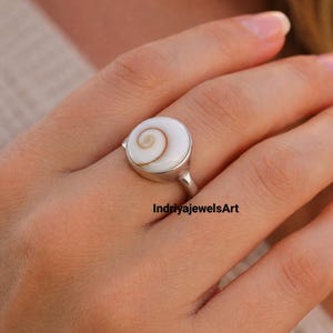 May include: A silver ring with a circular, white stone displaying a brown spiral design. The ring is on a finger. The text "IndriyajewelsArt" is visible.