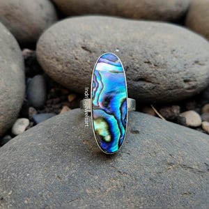 以下が含まれることがあります： 銀色のバンドが付いた、楕円形のアワビの貝殻の指輪。虹色の貝殻は、青、緑、紫の色合いの混合を示しています。指輪は灰色の石を背景に配置されており、指輪の色を際立たせています。