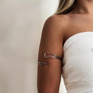 以下が含まれることがあります： ミニマリストデザインのシルバーカラーのアームカフ。カフは湾曲した上部と、端に小さなボールが付いたまっすぐなバーが特徴です。ジュエリーは上腕に着用され、モダンなエレガンスを添えています。