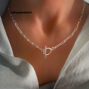 Pode incluir: Um colar de corrente prateada com elos retangulares alongados e um fecho de barra central. O colar está posicionado à volta do pescoço, com a barra inserida através de um anel circular. O texto indriyajewelsArt é visível.