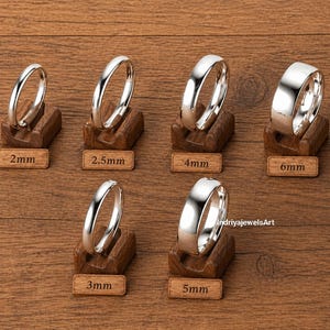 May include: A selection of silver rings, each presented on a small wooden stand. The rings are of varying widths, with labels showing sizes from 2mm to 6mm. The rings have a polished, reflective finish, set against a warm, wooden backdrop.