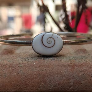 Może przedstawiać: Srebrna bransoletka z białą, owalną centralną częścią z brązowym wzorem spirali. Bransoletka jest umieszczona na teksturowanej, czerwonobrązowej powierzchni z rozmytym tłem.